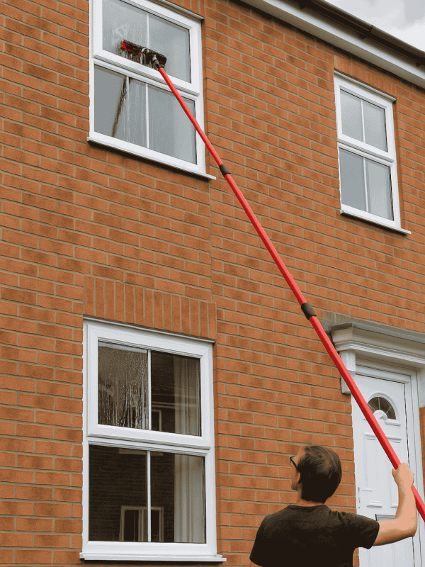 A person cleaning the windows of a building, wearing red clothing, outdoors using 4.6 water fed pole.